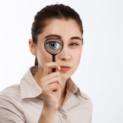 Young Beautiful Brunette Girl Looking With Mistrust At Camera Through Magnifier Over White Background. Young Beautiful Brunette Girl Looking With Mistrust At Camera Through Magnifier Over White Background.